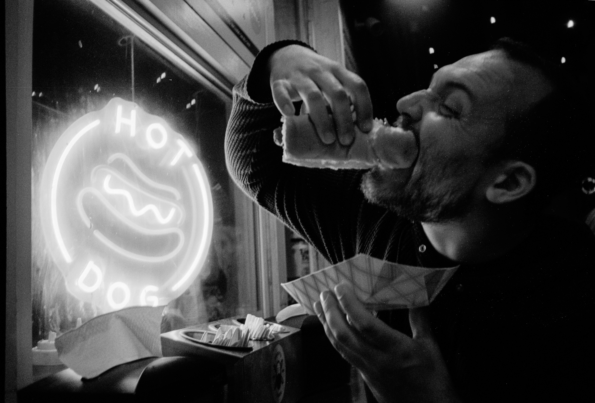 Black and white photo of a man eating a hot dog with Daddy's Dogs sign in the background.